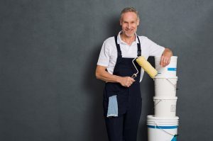 Man wearing white shirt and work apron holding paint roller and leaning a stack of painting supply buckets