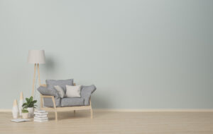 Unadorned interior wall with gray-green paint behind contemporary-style sitting room chair with darkish-green upholstery, eggshell-colored lamp, knickknacks and plantss